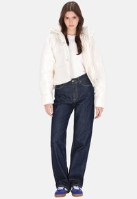 Hvid, cropped dunjakke med skinnende tekstur, kombineret med højtaljede mørkeblå jeans og blå sneakers. Modellen står mod en ensfarvet baggrund.