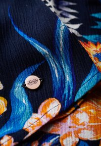 Il tessuto a motivo floreale presenta colori vivaci, tra cui blu, arancione e rosa. Un piccolo etichetta di marca è attaccata all'angolo.