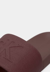 Sandale à glissière bourgogne avec une sangle texturée et un logo en relief. La semelle est plate et lisse, conçue pour un usage décontracté.