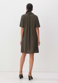 Olivenfarbenes Hemdkleid mit kurzen Ärmeln, Kragen und Rückenfalte. Der glatte Stoff endet über dem Knie und wird mit schwarzen, hohen Mules kombiniert.