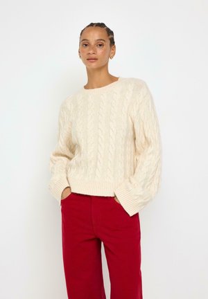 Kvinde med fletninger iført en cremefarvet kabelstrikket sweater og røde bukser, stående med hænderne i lommerne mod en hvid baggrund.