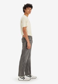Camiseta color beige claro, jeans rectos grises con un acabado desgastado, y zapatillas negras y blancas. El modelo está de perfil lateral contra un fondo blanco.