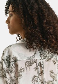 Leichte, durchsichtige Bluse aus Stoff mit Blumen- und Vogelmustern in Schwarz auf cremefarbenem Hintergrund, mit einer entspannten Silhouette und einem drapierten Ausschnitt.