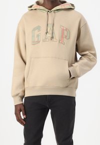 Beżowa bluza z kapturem z przednią kieszenią i sznurkiem do ściągania kaptura, z kolorowym logo "GAP" i kratkowaną podszewką wewnątrz kaptura.
