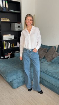 Γυναίκα στέκεται χαμογελαστή με λευκό πουκάμισο και μπλε φαρδιά τζιν παντελόνια σε ένα σαλόνι με μπλε καναπέ και μαύρα ράφια με βιβλία και αντικείμενα γραφείου.
