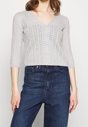 Kobieta ubrana w jasnoszarą, zapinaną na guziki kardigan z dekoltem w serek i wzorem w jodełkę oraz ciemnoniebieskie, wysokowalne dżinsy.