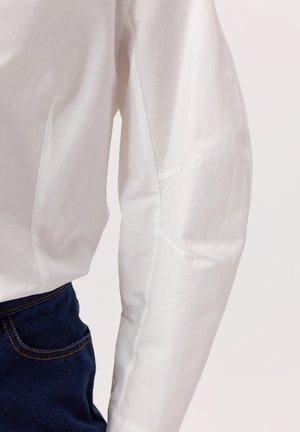 Biała bawełniana koszula o gładkiej fakturze, z dopasowanym rękawem i subtelnymi detalami szwów, w połączeniu z ciemnoniebieskimi dżinsami z pomarańczowym przeszyciem.