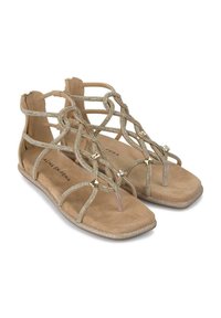 Sandalias planas beige con tiras metálicas entrelazadas, acentos de tachuelas y una suave plantilla de ante. Punta cuadrada y tacón mínimo.