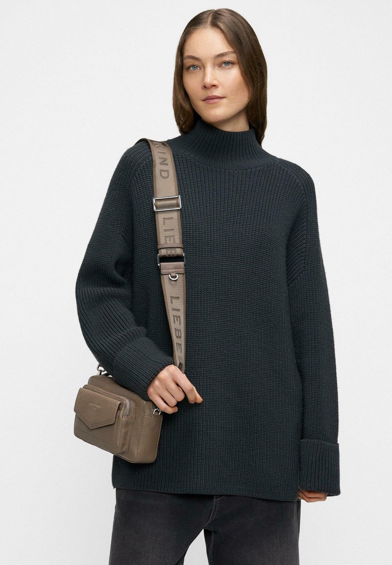 Kobieta ubrana w ciemnozielony prążkowany golf i czarne spodnie, niosąca małą torebkę na ramię w kolorze taupe z szerokim, oznakowanym paskiem.