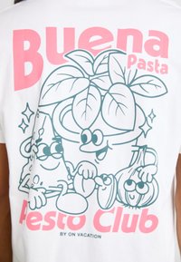 Graphic white t-shirt featuring a cartoon design of a plant, cheese, and garlic, with text "Buena Pasta" in pink and "Pesto Club" in blue.