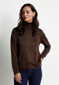 Pull à col roulé marron en matière douce, avec des manches longues et une coupe décontractée, associé à un jean bleu marine.