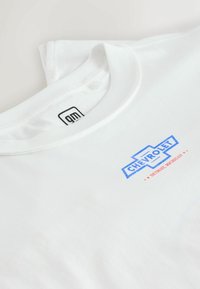 Tricou alb din bumbac, cu un logo Chevrolet albastru și roșu imprimat pe față. Guler rotund și detalii dintr-un material ribbed lângă guler.