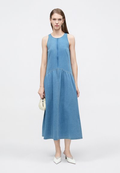 Γυναίκα που φοράει μπλε κοντομάνικο μίντι φόρεμα από τζιν, κρατάει μικρή κρεμ τσάντα χειρός και φοράει λευκές μυτερές γόβες με χαμηλό τακούνι, σε λευκό φόντο.