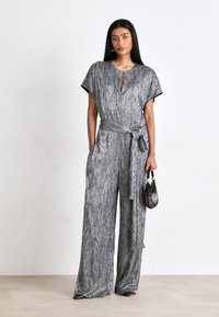 Sølvfarvet jumpsuit med mønstret, stribet tekstur, korte ærmer, nøglehulhalssnit og en bundet taljebælte. Stylized med en lille håndtaske.