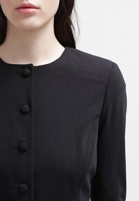 Svart klänning med hög halsringning, fem texturerade knäppknappar fram och långa ärmar i ett slätt, matt tyg.