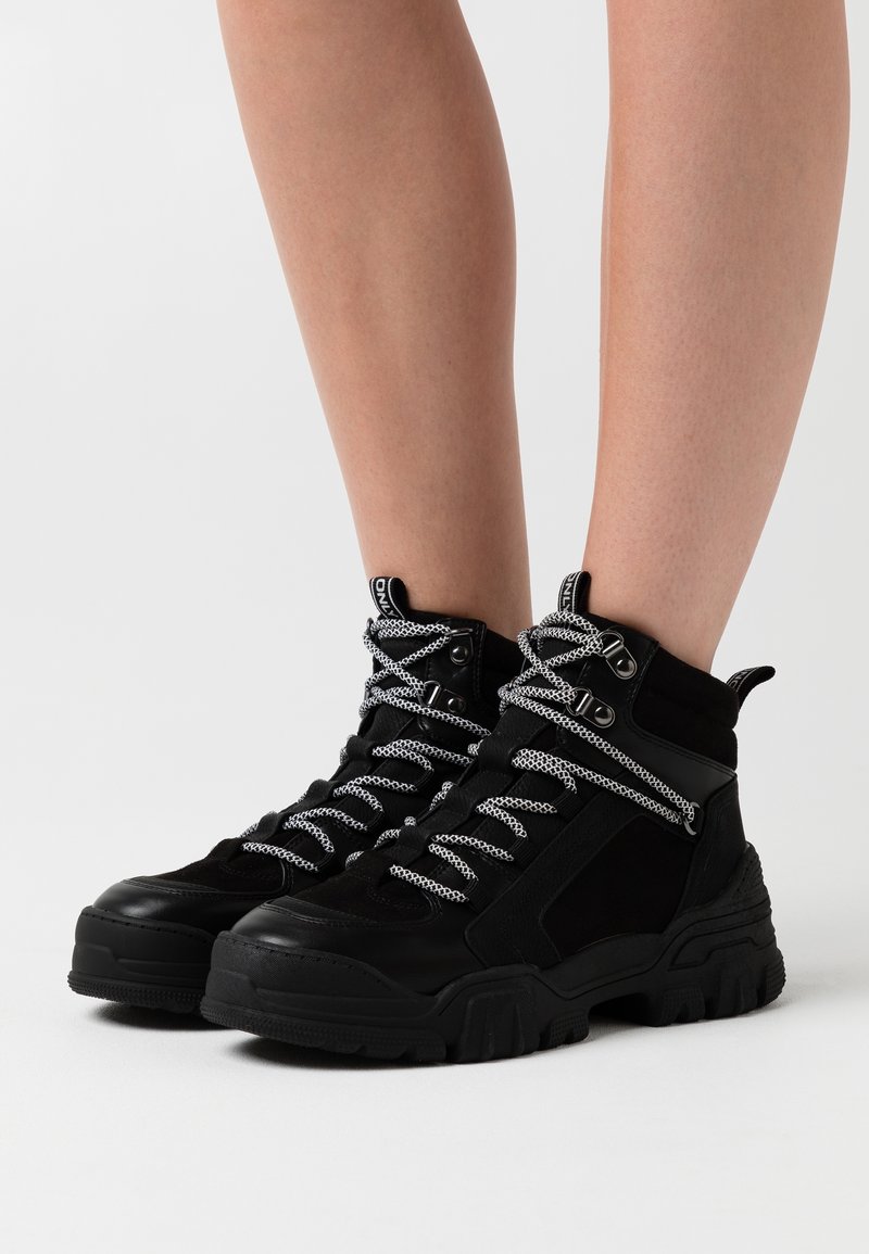 Bottes montantes noires épaisses avec des lacets épais à motifs portées sur des jambes nues, présentées sur fond blanc.