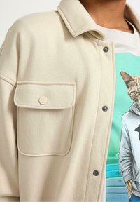 Camicia beige con bottoni, dotata di taschino sul petto, caratterizzata da una texture morbida e bottoni grigi a contrasto. Sotto, una t-shirt grafica con un design di un gatto.