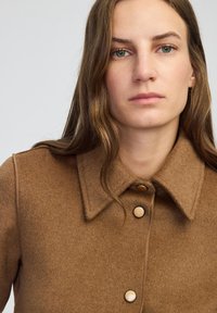 Brązowy wełniany płaszcz z kołnierzem, dwoma dużymi złotymi guzikami i teksturowanym wykończeniem. Prosty design z luźnym krojem.