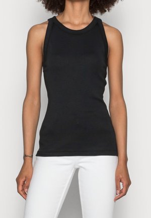 Persoon draagt een zwart mouwloos hemd en een witte broek, staand met ontspannen armen langs het lichaam tegen een effen achtergrond.