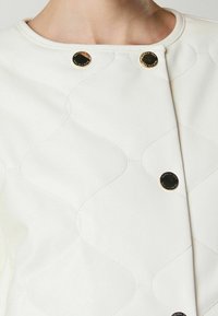 Giacca in tessuto bianco trapuntato con bottoni circolari dorati e dettagli di cucitura sottili, caratterizzata da un collo arrotondato e una texture liscia.