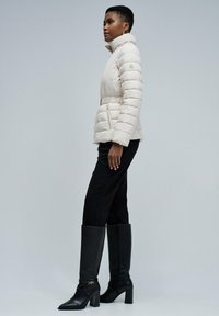 Doudoune crème avec coutures verticales, col montant, taille cintrée et poches latérales, portée avec un pantalon noir et des bottes noires montantes jusqu’au genou.