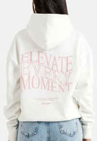 Felpa bianca con testo rosa sul retro che recita "ELEVATE EVERY MOMENT." Presenta un cappuccio con coulisse e polsini a costine. Materiale morbido, vestibilità comoda.