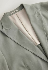 Blazer gris clair texturé avec revers crantés et doublure beige lisse, présenté à plat sur un fond clair.