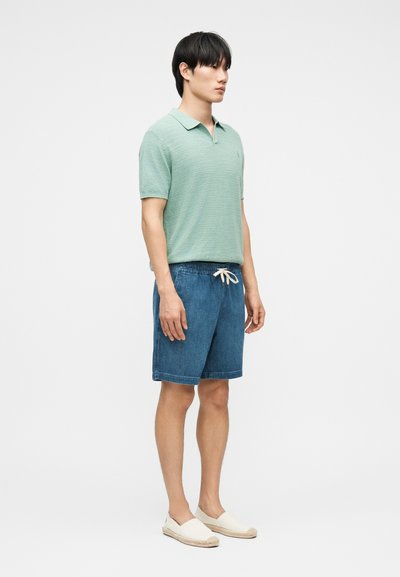 Νεαρός άνδρας που φοράει μπλουζάκι πόλο με κοντά μανίκια σε μέντα πράσινο χρώμα, μπλε τζιν σορτς με κορδόνι και κρεμ παπούτσια χωρίς κορδόνια, στέκεται πλαγίως.
