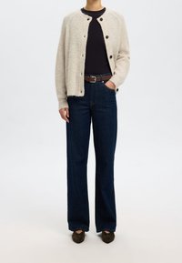 Ljus beige stickad kofta med rund hals och framknäppning, parat med mörkblå vida jeans och mörka skor.