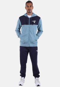 Felpa con cappuccio blu e navy con zip e coulisse, presenta un logo bianco sul petto. Abbinata a pantaloni della tuta navy e scarpe sportive bianche.