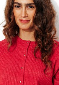 Cardigan rouge à côtes avec un col rond, arborant des boutons argentés sur le devant et un tissu texturé. Cheveux bouclés visibles.