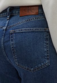 Tumed sinise tooniga teksapüksid, mis on hästi istuva lõikega. Omavad pruuni nahast vöökohatükki, millel on reljeefne logo, ning tagataskuid.