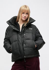 Sort dunjakke med høj krave, frontlynlås og trykknapper. Har quiltet design og broderet "Gap"-logo på brystet.