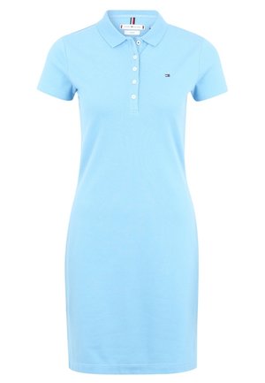 Kvinde iført en lyseblå polo-kjole med korte ærmer og hvide sneakers, stående med den ene arm bøjet og den anden afslappet.