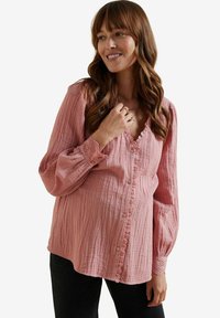 Blush-farbene Schwangerschaftsbluse, strukturierter Stoff, Rüschenausschnitt, Knopfverschluss, lange Ärmel mit Eyellet-Besatz an den Bündchen, getragen mit dunklen Hosen.