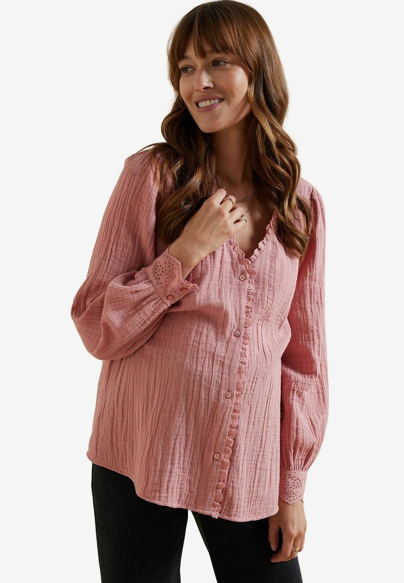 Blush-farbene Schwangerschaftsbluse, strukturierter Stoff, Rüschenausschnitt, Knopfverschluss, lange Ärmel mit Eyellet-Besatz an den Bündchen, getragen mit dunklen Hosen.