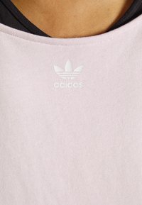 Camisa de algodão rosa clara com decote redondo, apresentando um logótipo branco da Adidas e uma textura com nervuras subtil. Tecido preto por baixo.