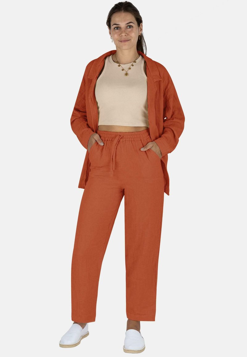 Ein leinenfarbener, rostfarbener Anzug, der ein lässig geschnittenes Hemd und eine Hose mit Kordelzug umfasst, kombiniert mit weißen Espadrilles. Glatte Textur und schlichtes Design.