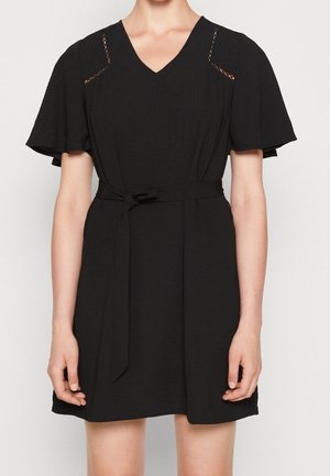 Woman wearing a short black dress with flutter sleeves, V-neck, lace accents on shoulders, and a tied fabric belt at the waist.