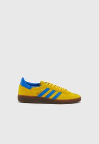Zapatilla de ante amarilla con acentos azules, que presenta tres rayas azules, una suela marrón texturizada y un diseño de cordones.