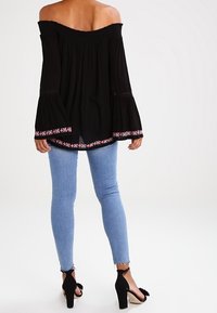 Donna che indossa un top nero scollato con ricami rosa e bianchi su maniche e orlo, jeans skinny blu chiaro e sandali neri con tacco alto.