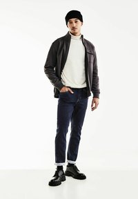 Conjunto masculino con una chaqueta de cerrar oscura, un suéter de cuello alto crema, jeans oscuros ajustados con puños y botas negras gruesas. Diseño sencillo y moderno.