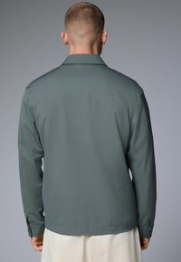 Chaqueta ligera de color verde oscuro con un diseño estructurado, mangas largas y un cuello clásico. Textura suave y puños abotonados.