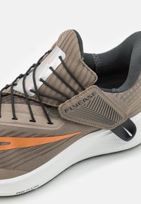 Lichtbruine sportschoen met een gestructureerde bovenkant, oranje accenten, FlyEase-technologie voor gemakkelijke toegang en een zwarte rubberen zool.