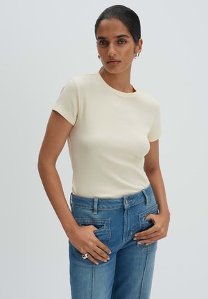 Donna con capelli scuri legati indietro che indossa una maglia crema a maniche corte a coste, jeans di jeans blu, orecchini a cerchio argento e anello d'argento, con le mani in tasca.