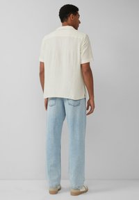 Korte mouwen, lichtcrème shirt met gestructureerde stof, gecombineerd met lichtblauwe, losse jeans en beige schoenen, gezien van achteren.