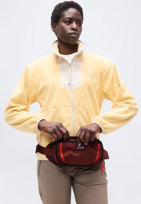 Άτομο που φοράει ανοιχτόκίτρινο φλις με φερμουάρ, μπεζ παντελόνια, προσαρμόζοντας μια μαρουν και κόκκινη τσάντα μέσης πάνω σε απλό λευκό φόντο.