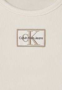 Tessuto bianco a coste con scollatura a girocollo presenta una toppa rettangolare con la scritta "Calvin Klein Jeans" ricamata in nero e oro.