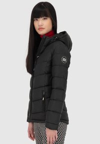 Giubbotto imbottito nero con cappuccio, caratterizzato da cuciture orizzontali, tasche con zip e una patch con logo sulla manica. Indossato con pantaloni a fantasia.