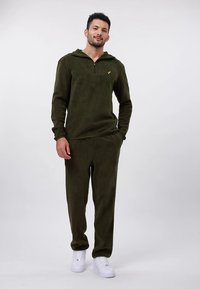 Olivově zelená fleecová tepláková souprava s kapucí na půl zipu. Kalhoty i vršek mají měkký vzhled, doplněné bílými teniskami.